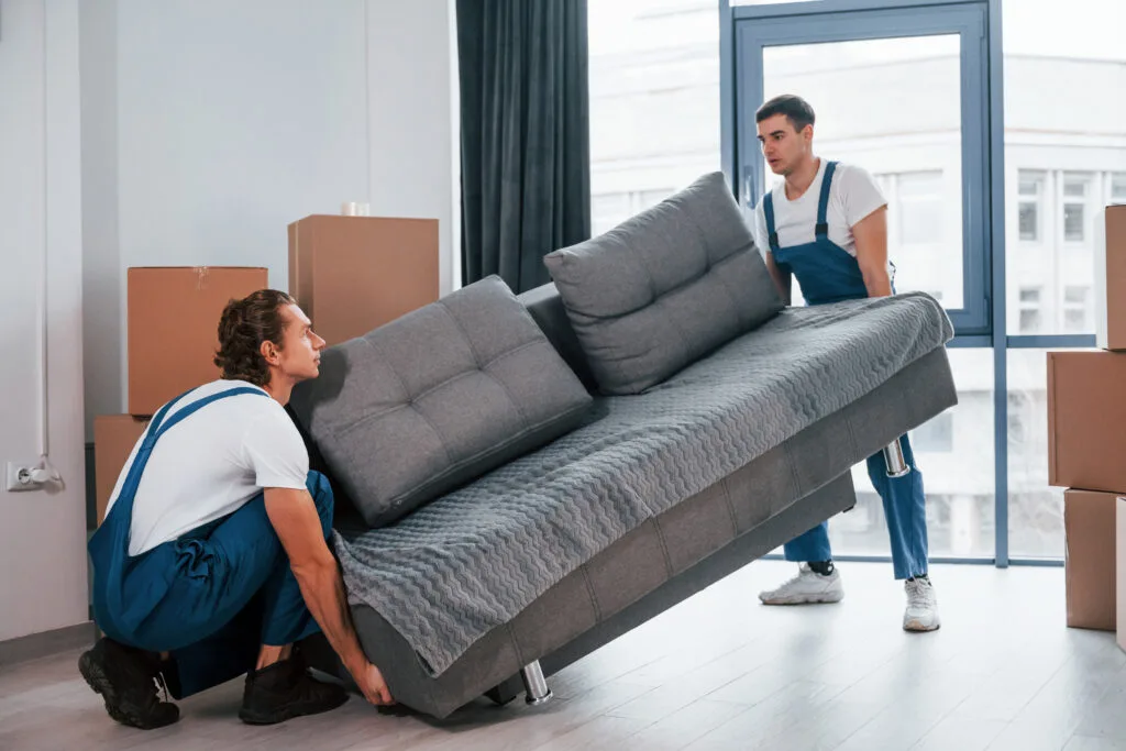 movers in ajman - Carrying heavy sofa. Two young movers in blue uniform working indoors in the room